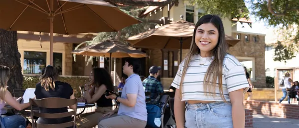 female student smiling on campus, with other students sitting at outdoor tables behind her working and chatting