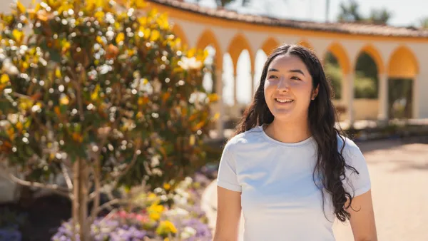 female student with long brown hair smiling walking outdoors with a corridor and some plants behind her