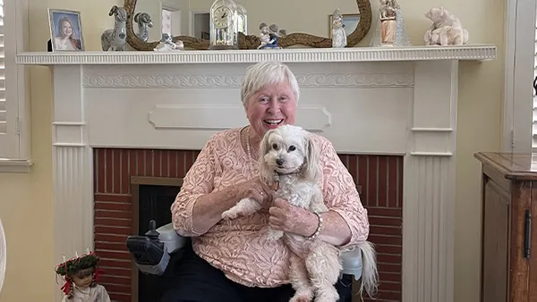 An older woman holding a small dog and smiling at the camera in a living room.