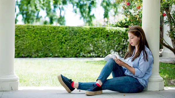 Young woman sitting and reading outdoors with a lawn and hedge in the background