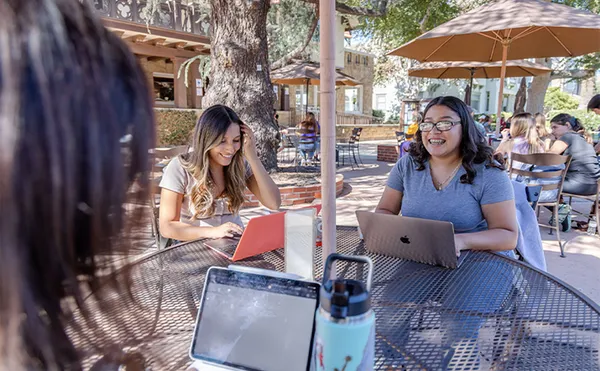 Three students in the foreground studying around a table under the shade of trees