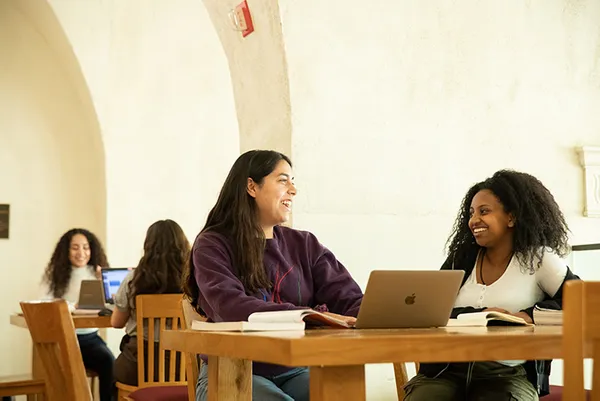students talking while sitting at a table with books and laptops, outdoors.