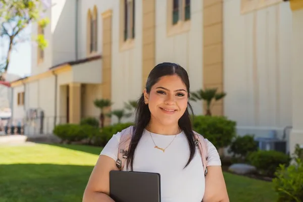 female student smiling standing outside of the chalon campus, holding a book and with a backpack on