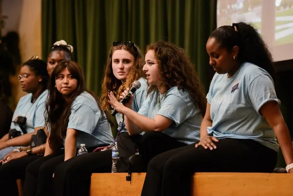 4 female students siting on stage in grey/blue shirts as one holds the mic and talks