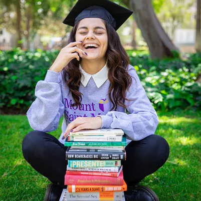 female student sitting on grass laughing with books in front of her