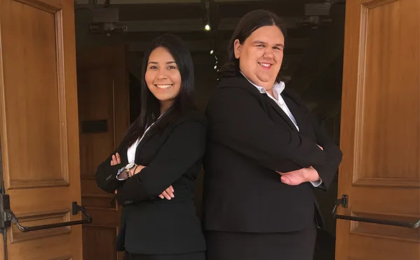Two women in business suits standing back to back and smiling at the camera.