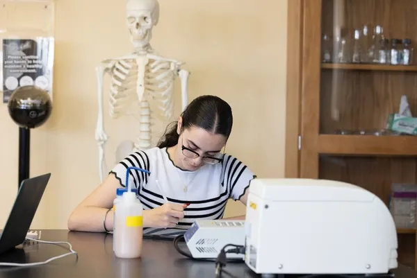 female student wearing glasses and a black and white shirt studying at a lab desk with a skeleton behind her