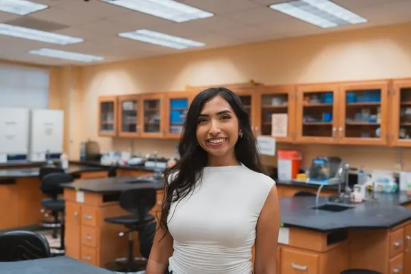 woman with long brown hair smiling wearing a white blouse standing in a classroom lab