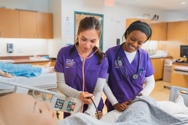 two female nursing students in purple msmu scrubs smiling as they work on a dummy patient in the nursing lab