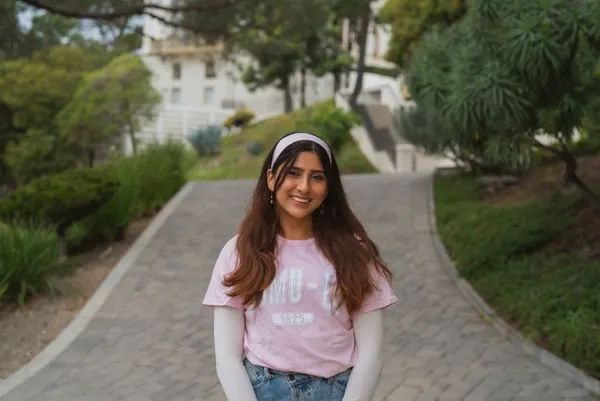 female student with brown long hair and white headband smiling outdoors in pink msmu shirt with white long sleeve shirt underneath and jeans