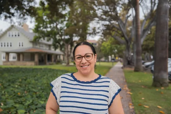 woman in striped blue and white shirt and wearing glasses smiling outdoors with trees and a building behind her