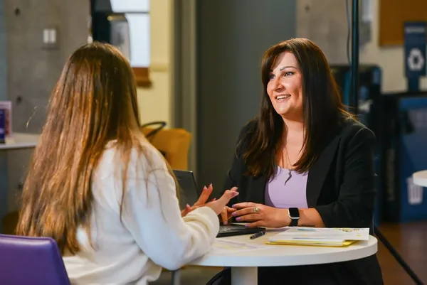 two women sitting at an indoor round table talking, one dressed professionally and listening to the other
