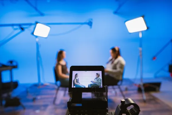 film set with a blue background and two women sitting facing each other talking, the foreground is focused on the camera that is pointed at them
