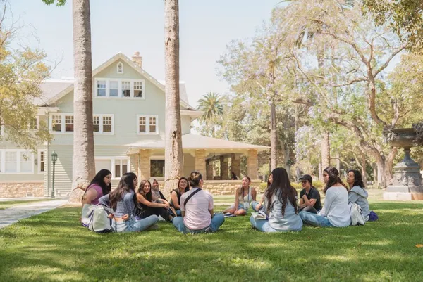 group of students sitting outdoors in a green lawn with a light green building in the background, sitting in a circle talking