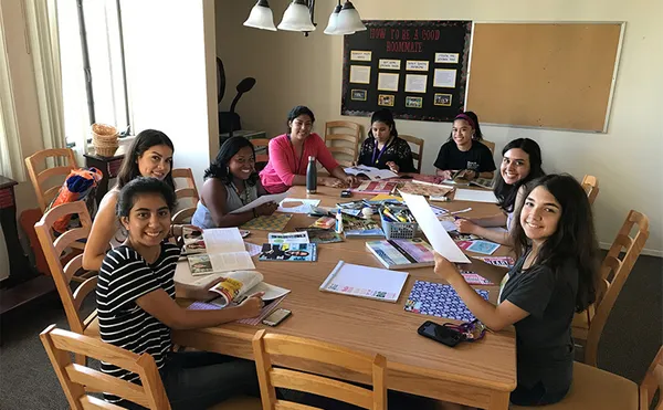 Students sitting around a table and smiling at the camera while lookng through magazines and papers.