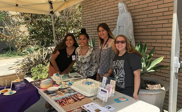 Four students smiling at the camera from behind a table covered with pamphlets and cake.