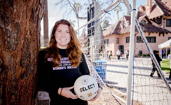 A student holding a soccer ball and leaning against a tree with a soccer goal and ˮ���� Doheny buildings in the background.
