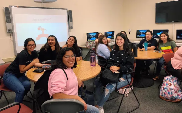 A group of students sitting around tables in a classroom and smiling at the camera