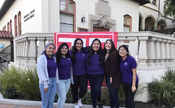 Six students smiling in front of a flag drapped over a columned railing