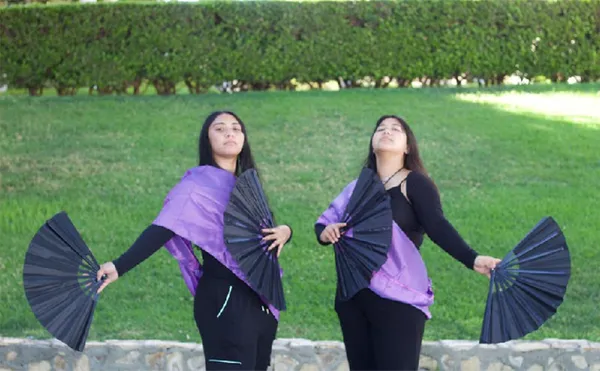 Two women holding folding fans and posing in front of a lawn and low hedge