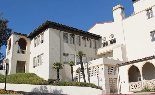 A large multi-level white building with a small driveway wrapping around lined with hedges.