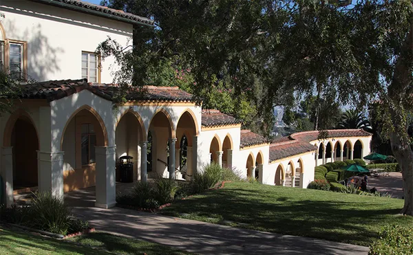 A building connected to an arched walkway which runs along an open central area with trees, hedges and tables.