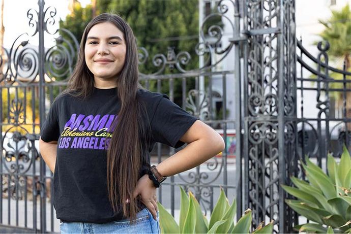 Female student smiling at front entrance to campus with an 91ɬ�� shirt on.