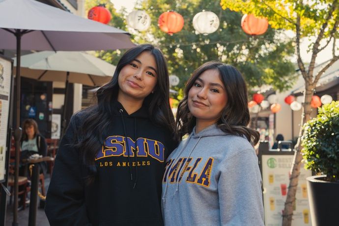 two female students standing together, one in a black 91ɬ�� sweater and one in a grey 91ɬ�� sweater.