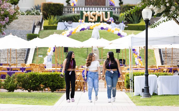 Three students walking along a paved path with tables, umbrellas and balloons in the background.